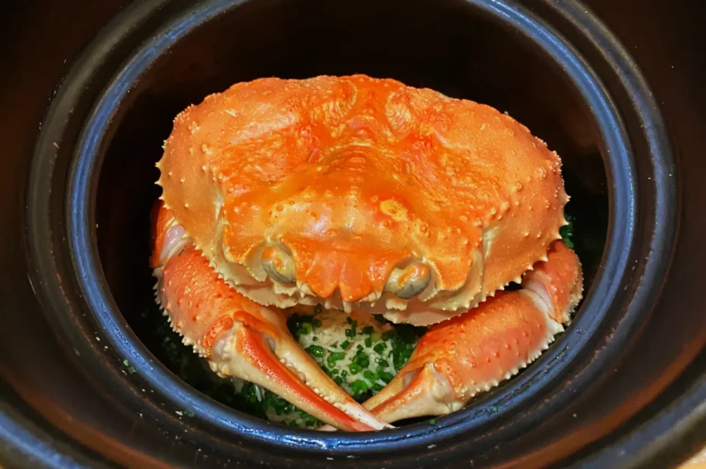 A vibrant orange crab sits in a black pot atop green diced herbs. The crab's claws and texture suggest it's ready for cooking, conveying a rustic culinary scene.