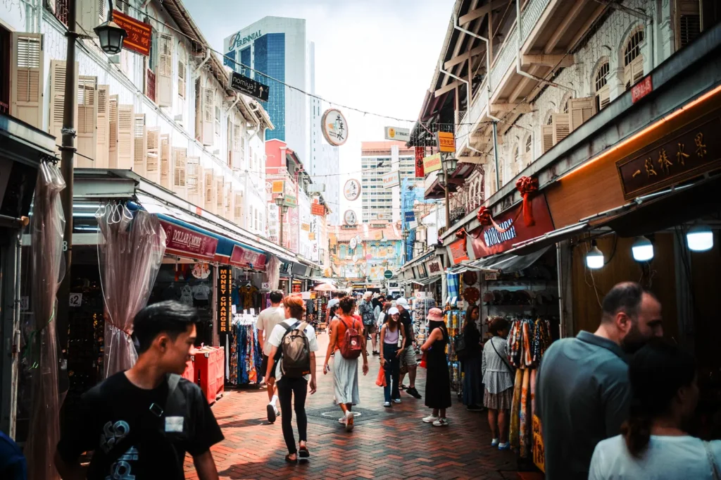 A bustling pedestrian street in Singapore’s Chinatown is lined with historic multi-story shophouses featuring louvered windows and street-level souvenir shops. Numerous locals and tourists walk along the brick-paved path under a string of circular lanterns, with modern city high-rises visible in the background.