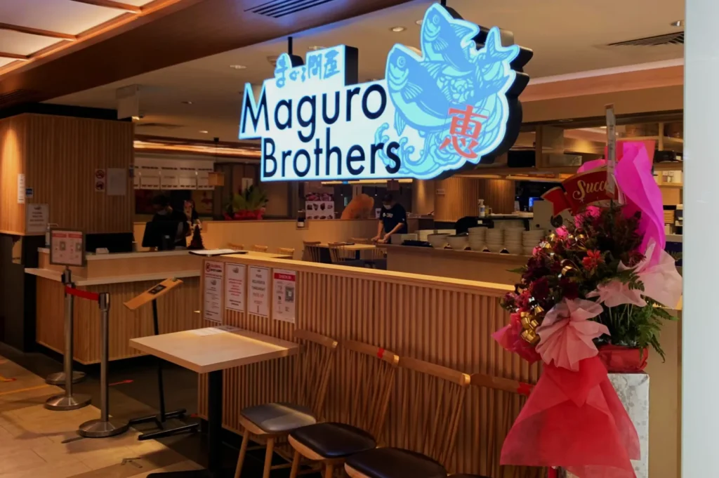 Restaurant entrance of Maguro Brothers, featuring a bright sign with stylized graphics. Seating area includes a floral arrangement, creating a welcoming atmosphere.
