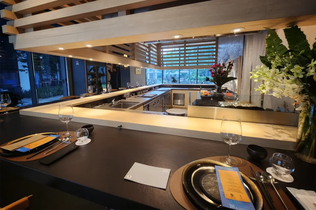 A wide-angle, eye-level shot captures a sleek, dark dining counter meticulously set with formal dinnerware, menus, and wine glasses for guests. The view looks across into a pristine, open U-shaped kitchen station featuring a warmly illuminated service ledge, stainless steel appliances, and fresh floral arrangements under a slatted wood ceiling structure.
