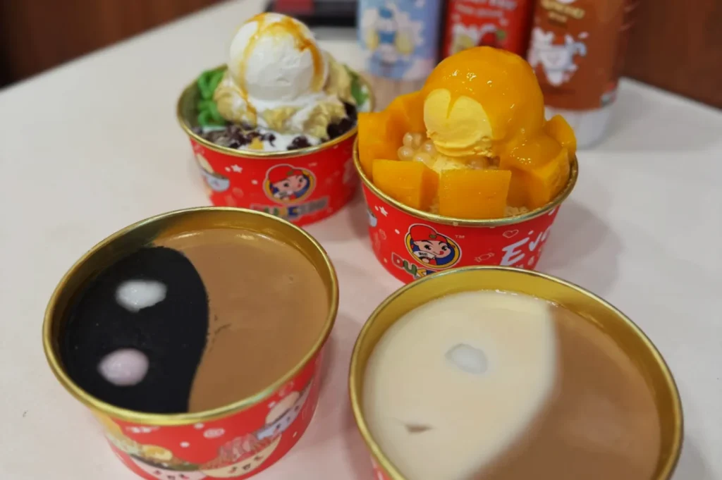 Four colorful dessert bowls on a table. Two show yin-yang designs, while others have ice cream with greens, red beans, and mango cubes.