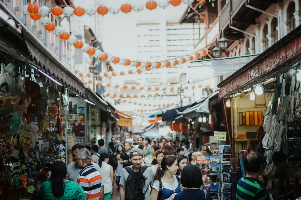 A high-angle shot captures a dense crowd of shoppers walking down a narrow street market lined with souvenir stalls under strings of hanging red and white lanterns. The bustling pathway is framed by traditional multi-story buildings, with a modern white high-rise building visible in the soft-focus background.