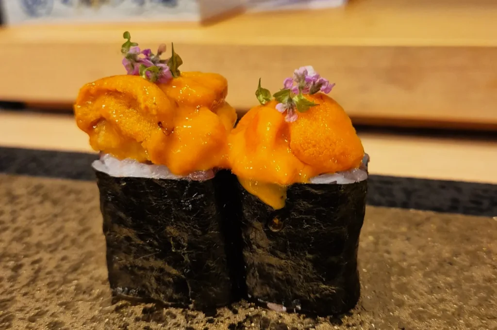 Two pieces of uni sushi with vibrant orange sea urchin on rice wrapped in nori, garnished with delicate purple flowers, set on a textured plate.