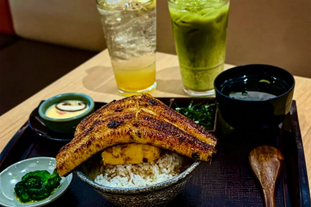 A high-angle shot captures a Japanese unagi set meal served on a dark tray, featuring a large, glazed grilled eel fillet resting on top of tamagoyaki egg and white rice. The meal includes sides of chawanmushi, clear soup, pickled greens, and a wooden spoon, with two tall iced drinks visible in the background.