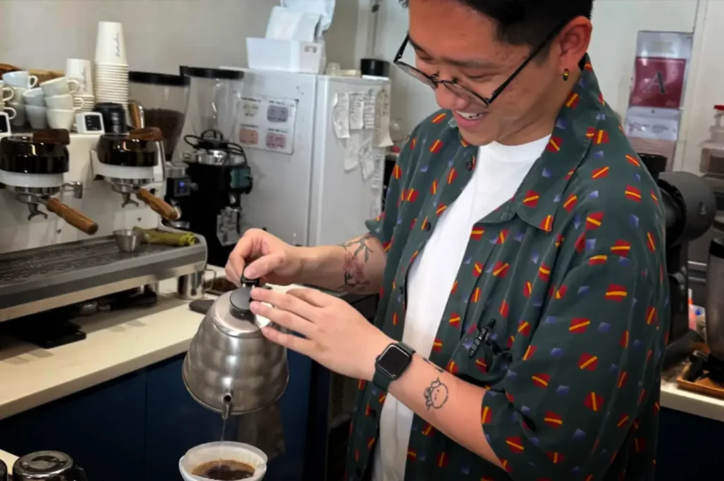 An eye-level, medium close-up shot captures a smiling barista pouring hot water from a silver, ridged gooseneck kettle into a pour-over coffee dripper. The barista, wearing glasses, an open green button-up shirt with geometric patterns, and a white t-shirt, stands on the right side of the frame with visible tattoos on his forearm. In the background on the left, a professional espresso machine sits on a counter alongside stacks of white cups, paper cups, and coffee grinders, while a white refrigerator and kitchen equipment complete the busy coffee shop interior.