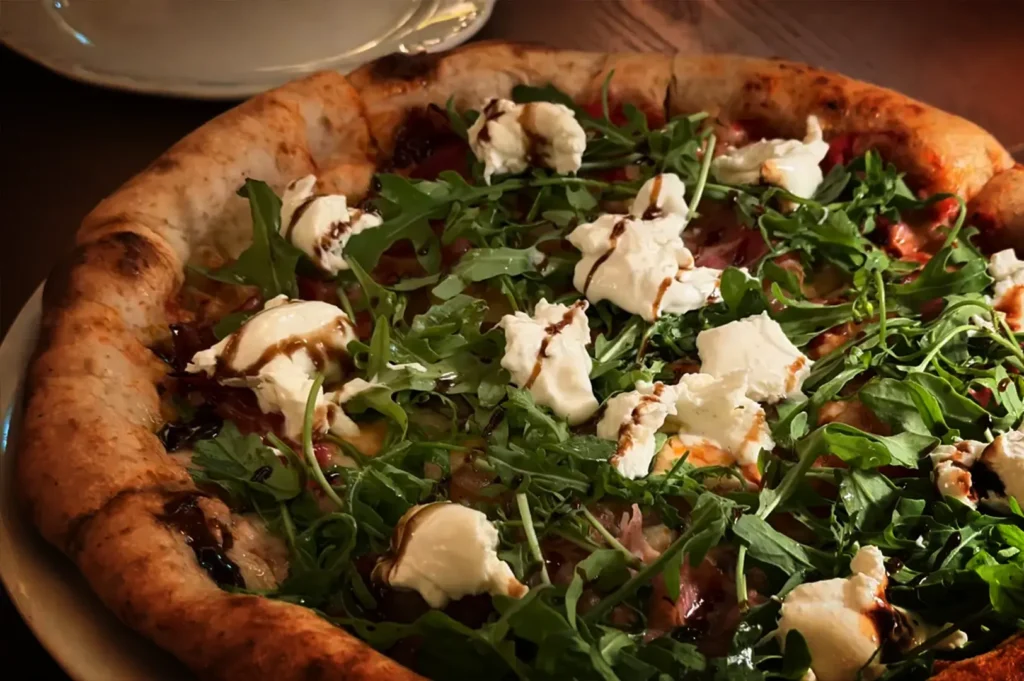 A close-up, slightly high-angle shot captures a freshly baked artisanal pizza served on a white plate over a wooden table. The pizza features a thick, golden-brown, puffed crust framing a bed of vibrant green arugula leaves. Scattered generously across the greens are dollops of soft, white burrata or mozzarella cheese, which are drizzled with a dark, glossy balsamic glaze. A hint of cured meat or prosciutto is visible beneath the toppings, and the stem of a wine glass is partially seen in the upper right background.