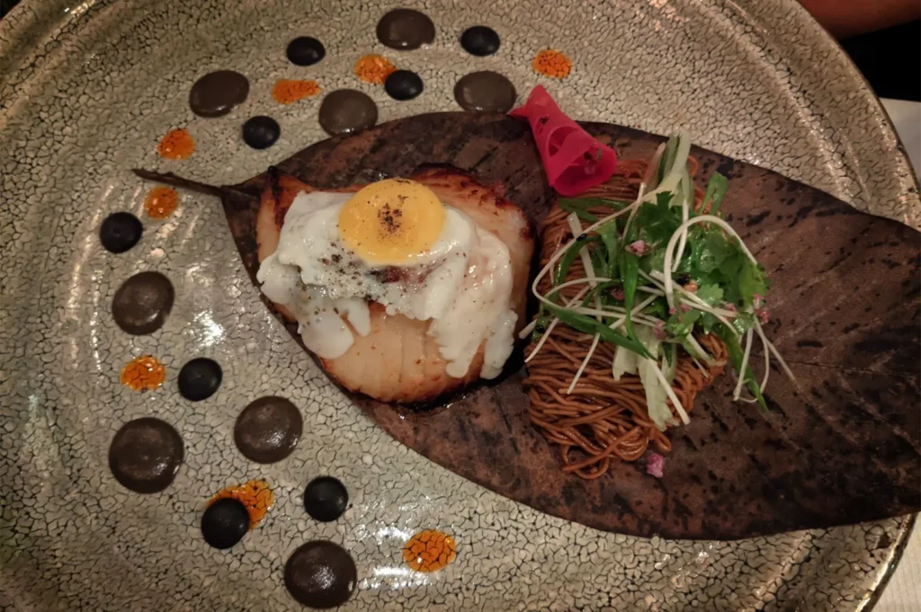A high-angle, close-up shot captures an exquisitely plated entrée presented on a textured, crackle-glazed ceramic plate. Resting at the center on a large, dried brown leaf is a pan-seared piece of white fish topped with a small, perfectly fried quail egg seasoned with black pepper. To the right of the fish sits a neat bundle of thin, brown noodles garnished with a fresh salad of julienned cucumbers, scallions, and cilantro, alongside a bright pink pickled accent. The surrounding plate surface is artistically decorated with alternating drops of smooth black and textured orange purées, completing the sophisticated culinary presentation.