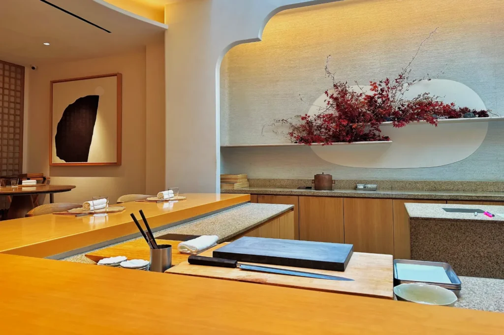 Minimalist sushi bar interior with a wooden counter set for guests. A knife and plate are on display, with red floral arrangement and abstract art adding elegance.