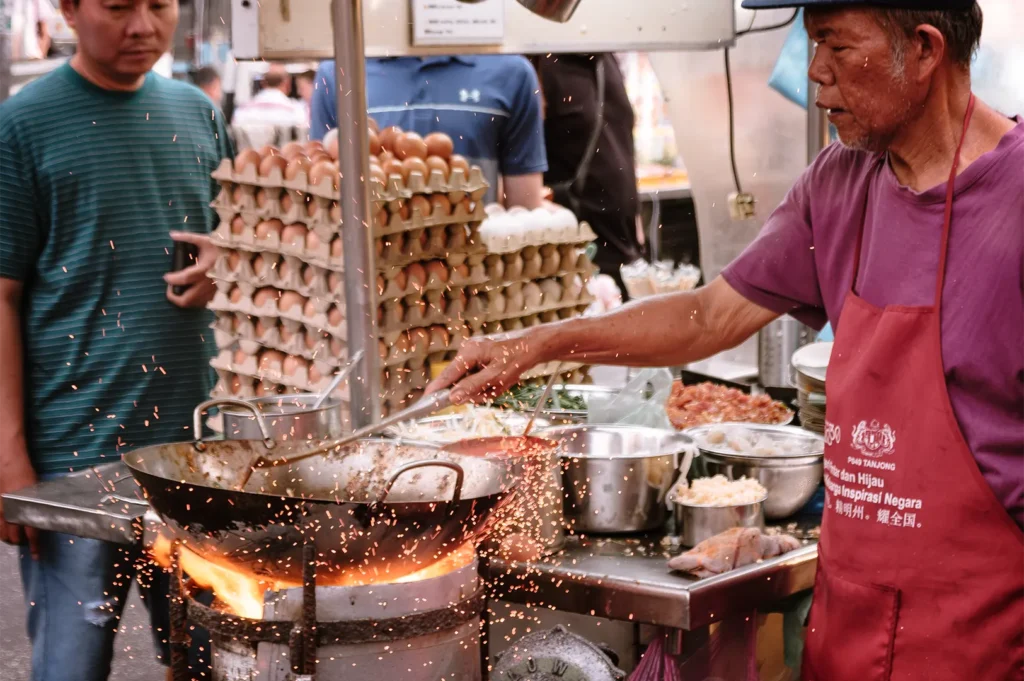 An eye-level shot captures a street food vendor tossing ingredients in a large wok over a powerful open flame, sending bright sparks flying into the air. Beside his stainless steel workstation, a tall stack of egg cartons and various ingredient bowls are visible as a patron looks on.