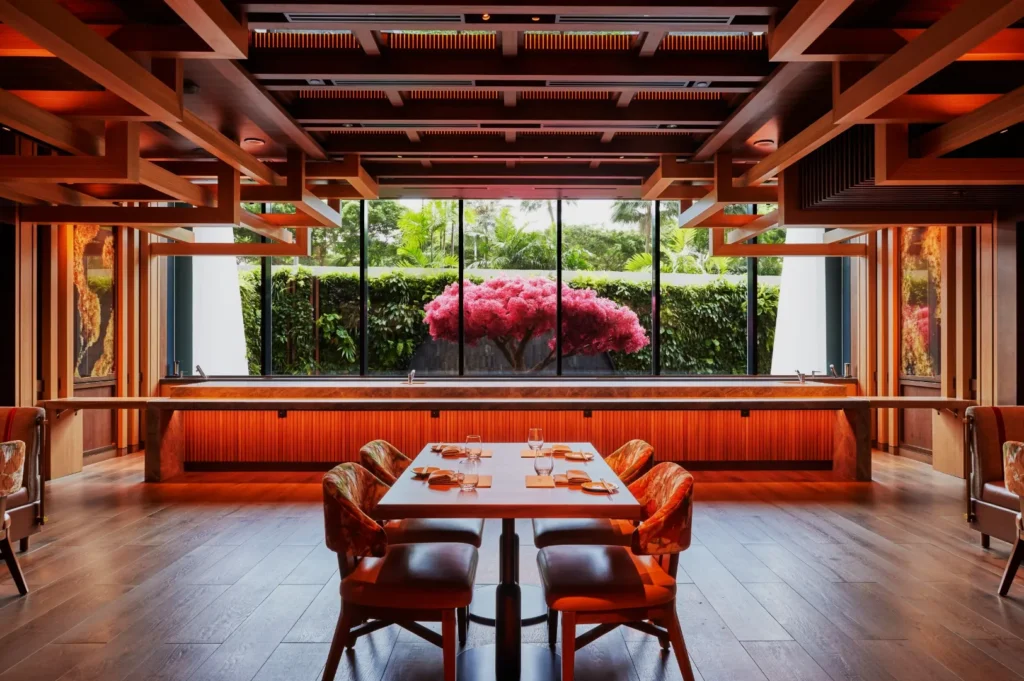 Sophisticated restaurant interior with wooden decor and tables set for dining. Large window showcases a lush garden and vibrant pink tree, creating a serene ambiance.
