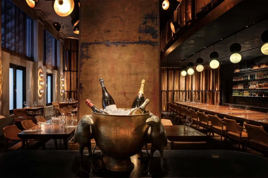 An eye-level shot captures a large brass ice bucket engraved with "EVERMORE" holding chilled wine bottles, flanked by two distinctive bird sculptures in the foreground. The moody, upscale restaurant interior features warm, artistic lighting, textured walls, and long wooden dining tables lined with leather chairs.