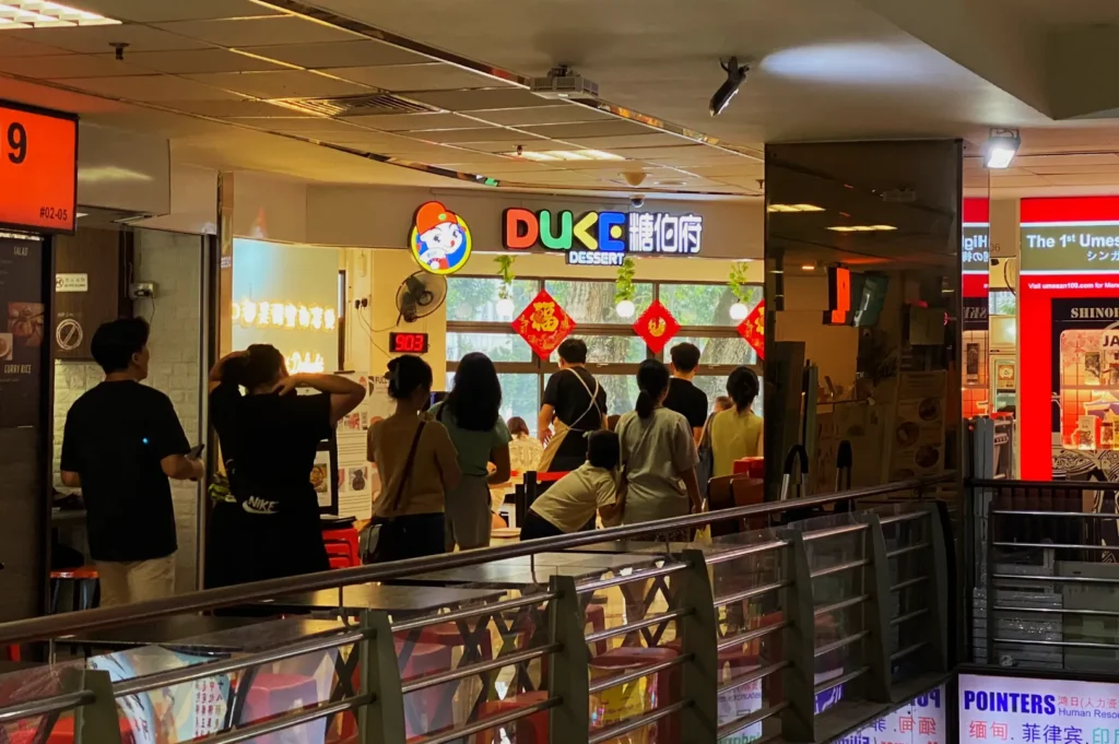 People stand in line outside "DUKE Dessert" in a mall. The bright neon sign and festive red decorations create a lively atmosphere.