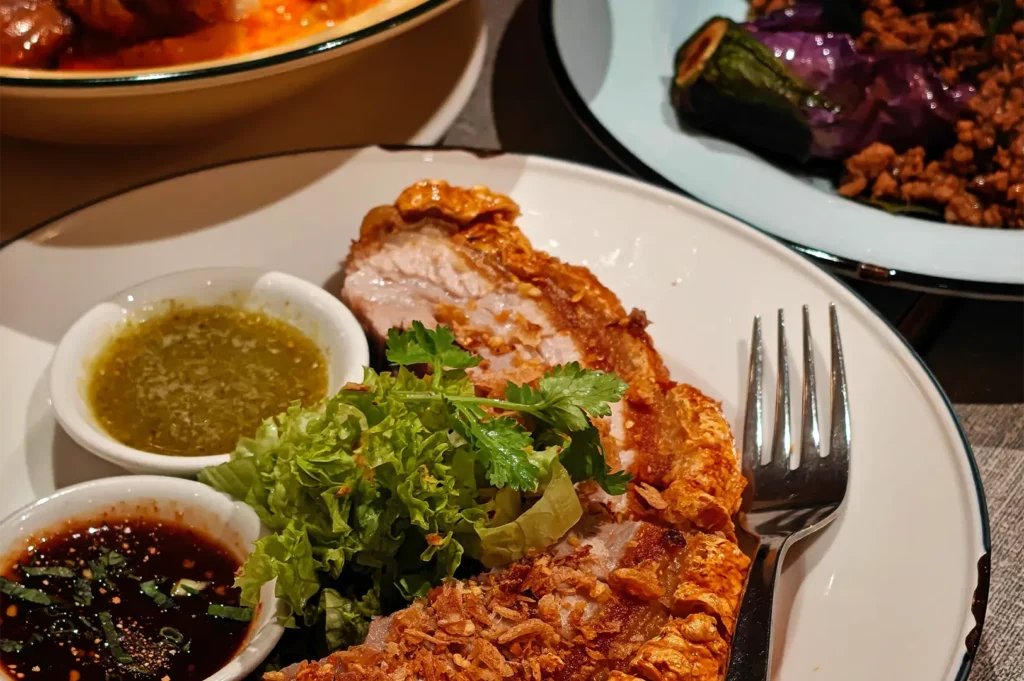A high-angle, close-up shot focuses on a white ceramic plate featuring crispy roast pork belly with a golden, crackled skin, garnished with fresh lettuce and cilantro. The dish is served alongside two small dipping sauces—one dark and one green—with a silver fork resting on the side of the plate.