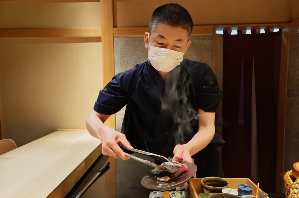 Chef in a mask carefully plates steaming food with tongs, wearing a dark uniform. The setting is a minimalist kitchen with warm lighting, conveying focus and precision.