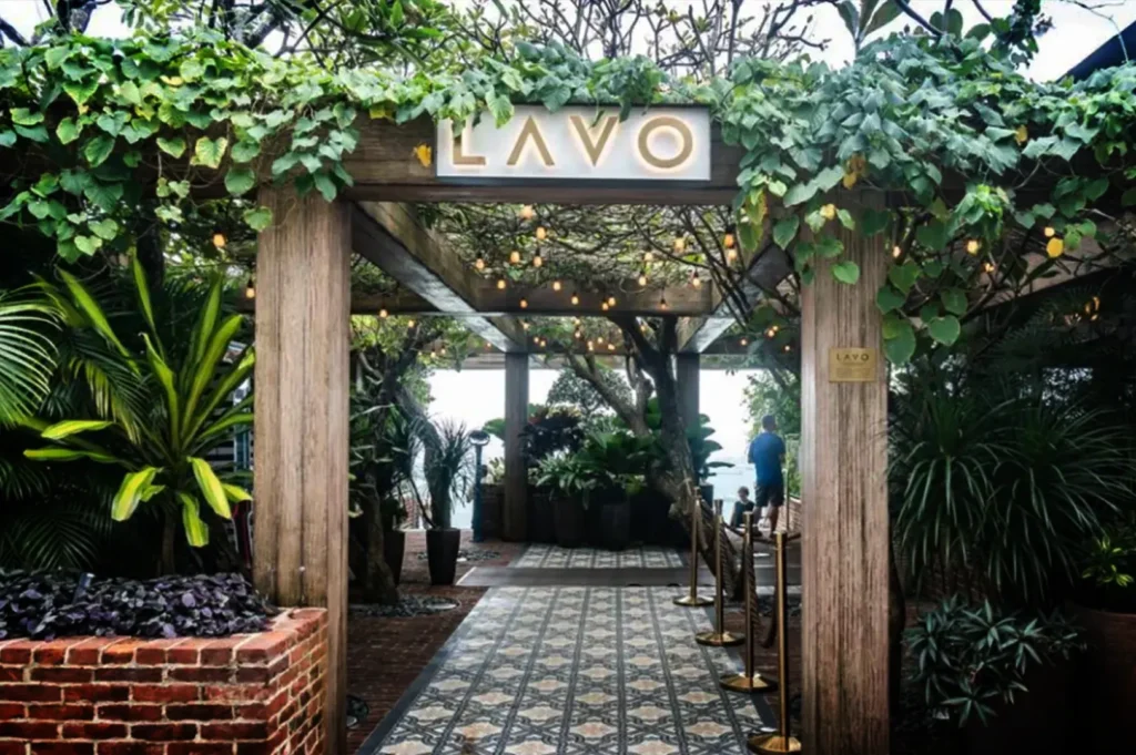 An eye-level, straight-on shot captures the lush, garden-like entrance to the restaurant LAVO. Thick wooden pillars and an overhead trellis support a dense canopy of green vines and hanging string lights, with a sleek white sign displaying the backlit gold text "LAVO" mounted at the center of the entryway. A decorative, patterned tiled walkway leads under the trellis toward an open-air terrace filled with potted plants and trees, where a person in a blue shirt is visible looking out toward the distance. The entrance is framed by vibrant tropical greenery, a low red brick planter box on the left, and large potted shrubs on the right, creating a warm and inviting atmosphere.