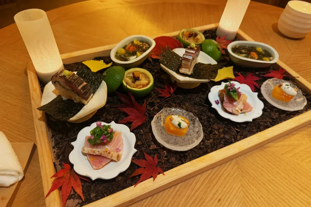 A wooden tray with assorted appetizers, including slices of fish, seaweed, and fruit. Garnished with colorful leaves and arranged for fine dining.