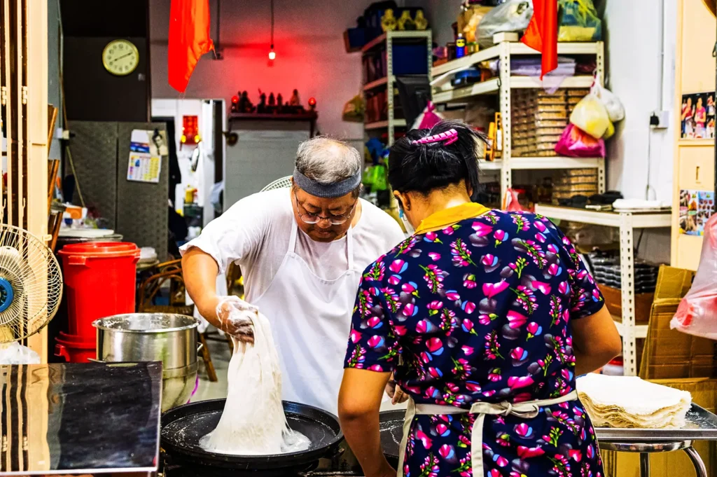 An eye-level shot captures a vendor in a white apron pulling and stretching a large mound of white dough over a flat circular griddle. Beside him, a woman in a vibrant floral shirt works at a separate station next to a neat stack of freshly made, thin pastry sheets in a busy commercial kitchen.