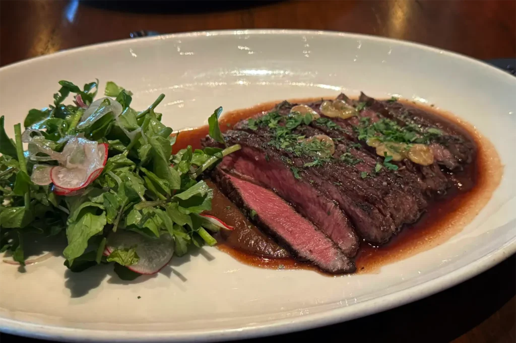 A close-up, eye-level shot highlights a perfectly sliced medium-rare steak resting in a rich, dark pan sauce, garnished with chopped herbs and roasted garlic chips. The main course is served on an oval white plate alongside a fresh green salad tossed with thinly sliced radishes.