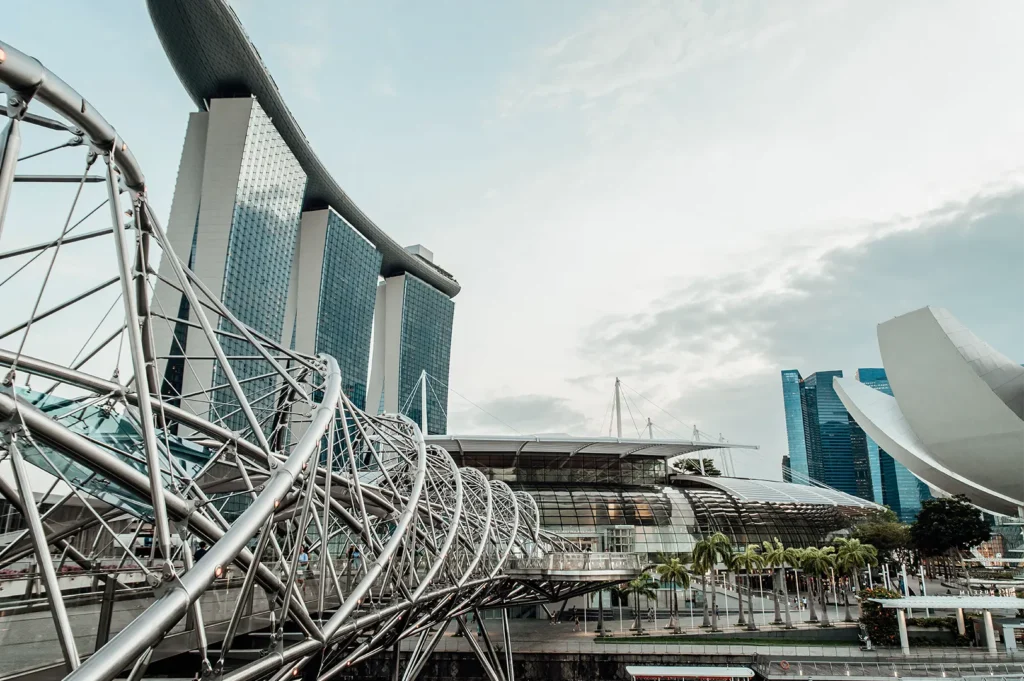 A low-angle shot features the intricate, double-helix stainless steel structure of the Helix Bridge stretching across the left foreground. In the background, the soaring towers of the Marina Bay Sands resort and the white, petal-like structure of the ArtScience Museum stand under a bright, overcast sky.
