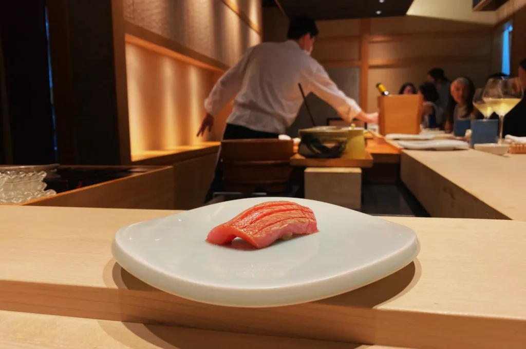 Sushi on a white plate is in focus at a minimalist sushi bar. A chef is preparing food in the background, while seated patrons watch attentively.