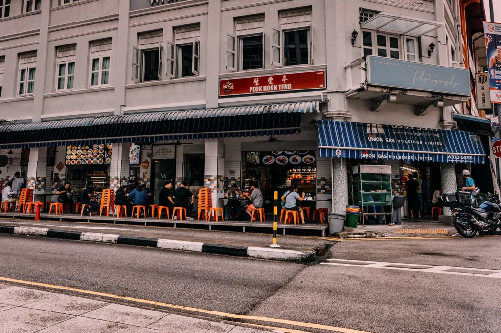 An eye-level shot captures a row of traditional street-level food stalls under blue-and-white striped awnings on a city corner building. Patrons sit on orange plastic stools at outdoor tables enjoying meals, while a parked motorcycle stands near the crosswalk in the foreground.